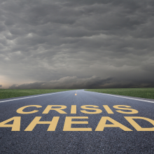 Road with the words "Crisis Ahead" painted on the ground in yellow paint. Storm clouds are visible in the background.