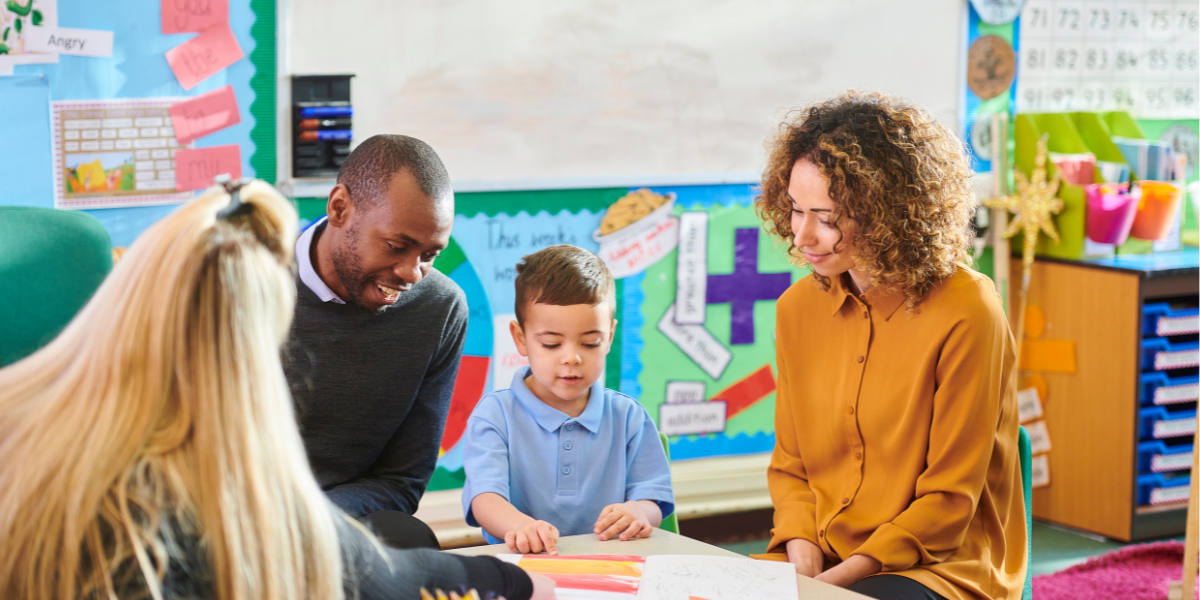 A teacher points to a book as a young child reads between his parents.