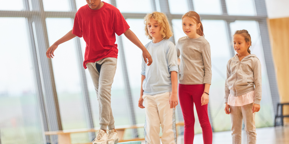 Child in a red sweatshirt walks on a balance beam as three classmates look on.