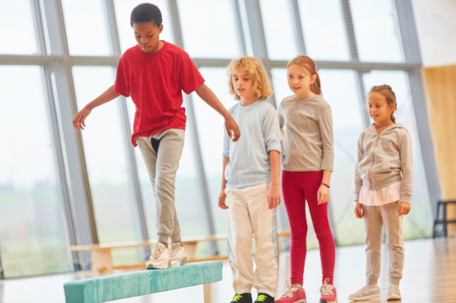 Child in a red sweatshirt walks on a balance beam as three classmates look on.