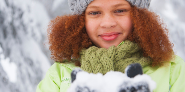 A child with curly red hair wearing a toboggan, smiles while holding a snow outside.