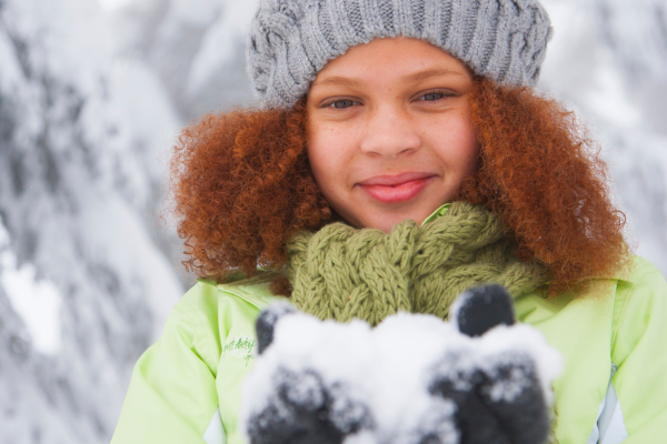 A child with curly red hair wearing a toboggan, smiles while holding a snow outside.