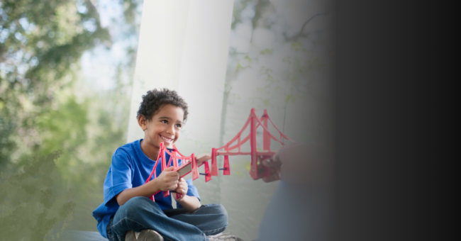 Child wearing a blue shirt and blue jeans playing with a red toy bridge